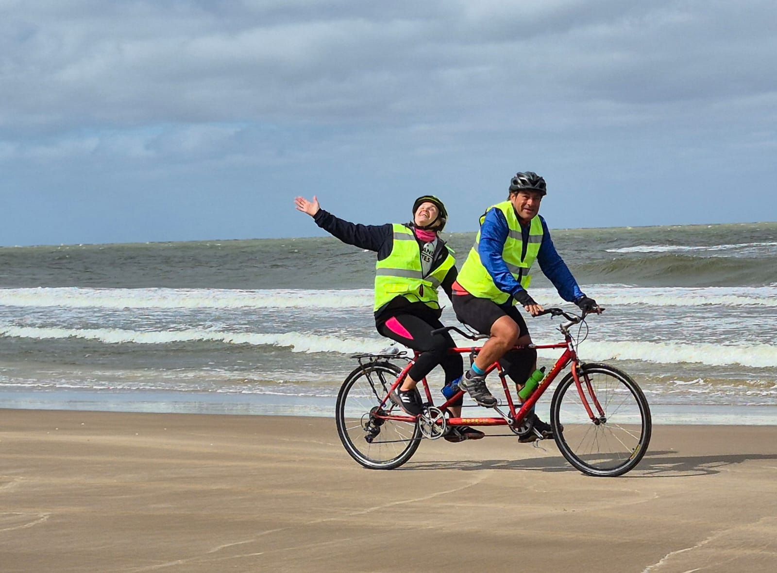 Dos integrantes de Atreverse en bicicleta tándem sobre la arena de la playa: quien va adelante conduce y quien va atrás levanta los brazos en señal de alegría