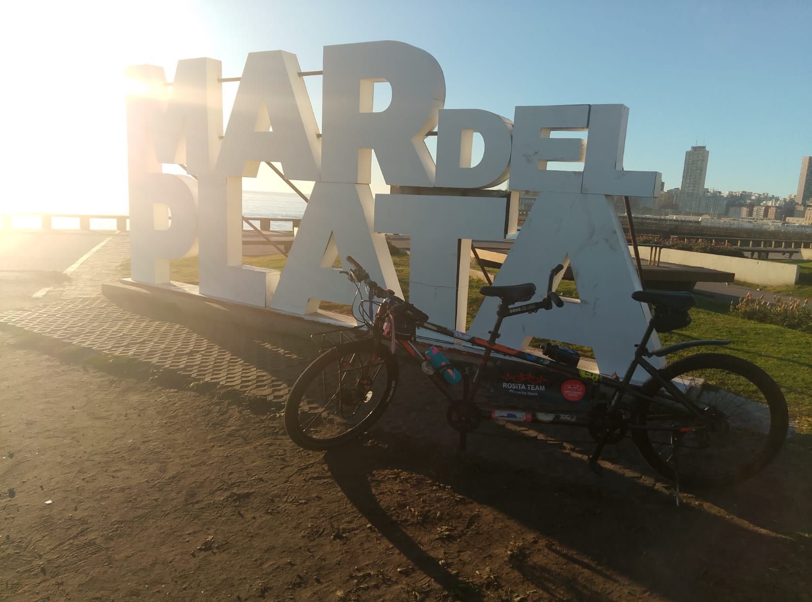 Cartel blanco con la inscripción MAR DEL PLATA junto al mar, con una bicicleta tándem adornada con calcomanías de Rosita Team en primer plano
