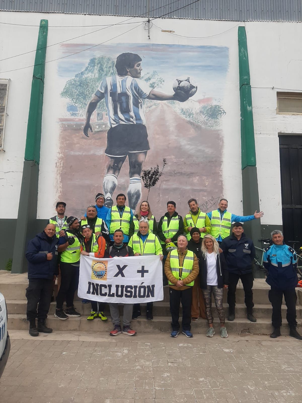 Grupo Atreverse frente a un mural de Maradona sosteniendo una bandera que dice Por más INCLUSIÓN