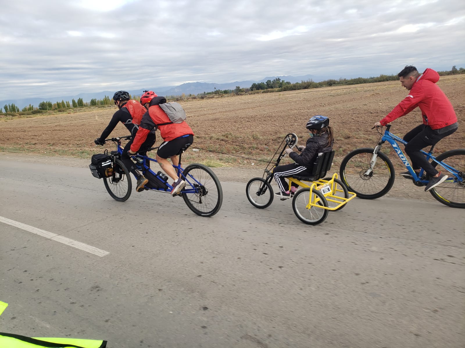 Cuatro personas pedaleando en un entorno rural: dos adultos en bicicleta tándem y una niña en bicicleta adaptada, acompañados por un ciclista guía